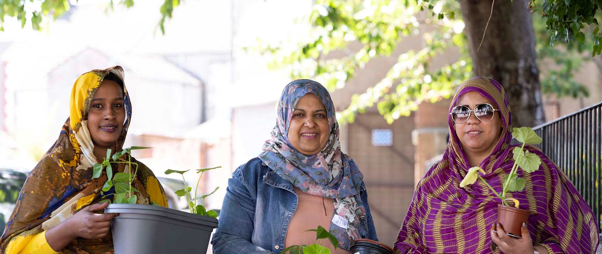 Three women wearing headscarves holding plants in pots and smiling